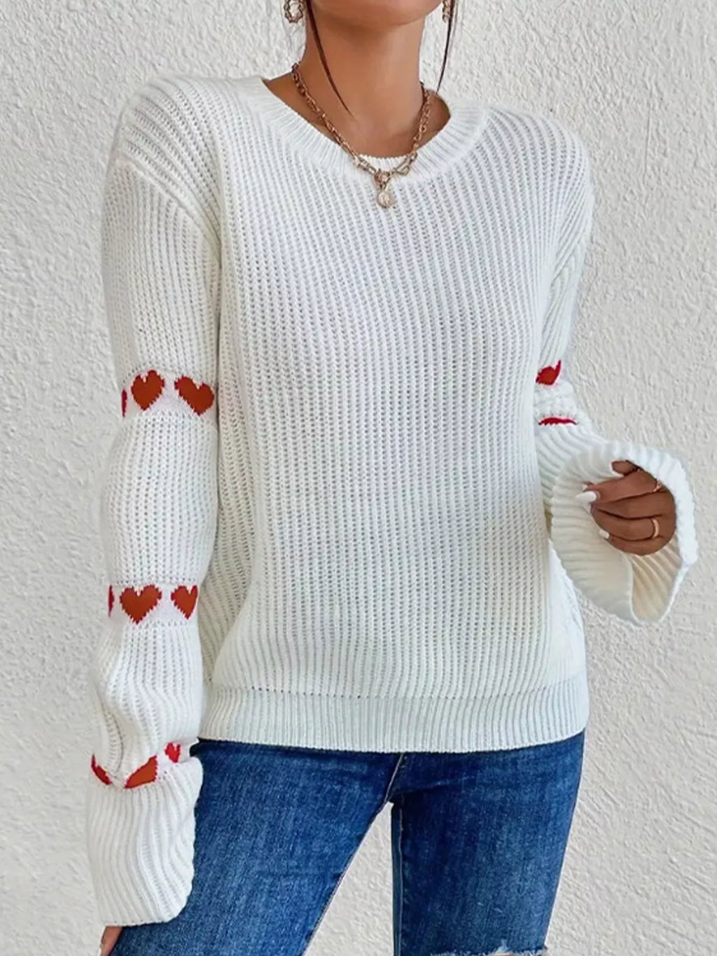 Pull blanc en maille à col rond et cœurs rouges sur les manches, porté avec un jean bleu.