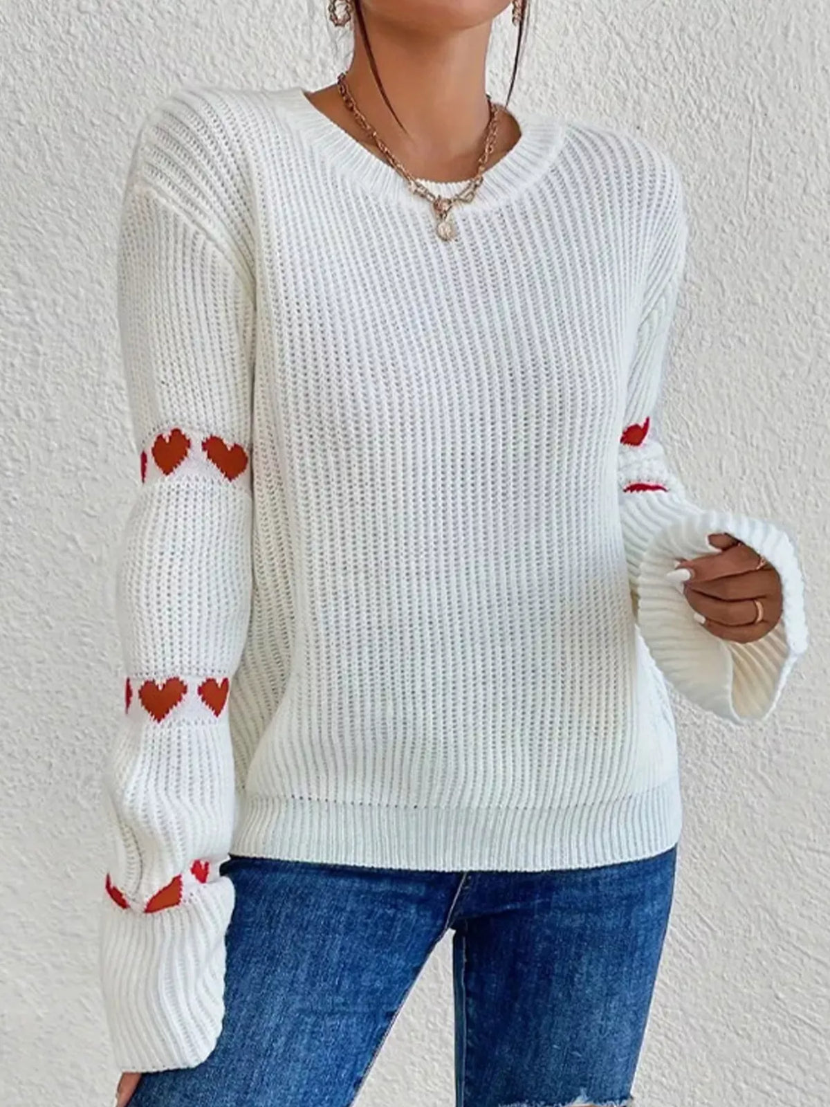 Pull blanc en maille à col rond et cœurs rouges sur les manches, porté avec un jean bleu.